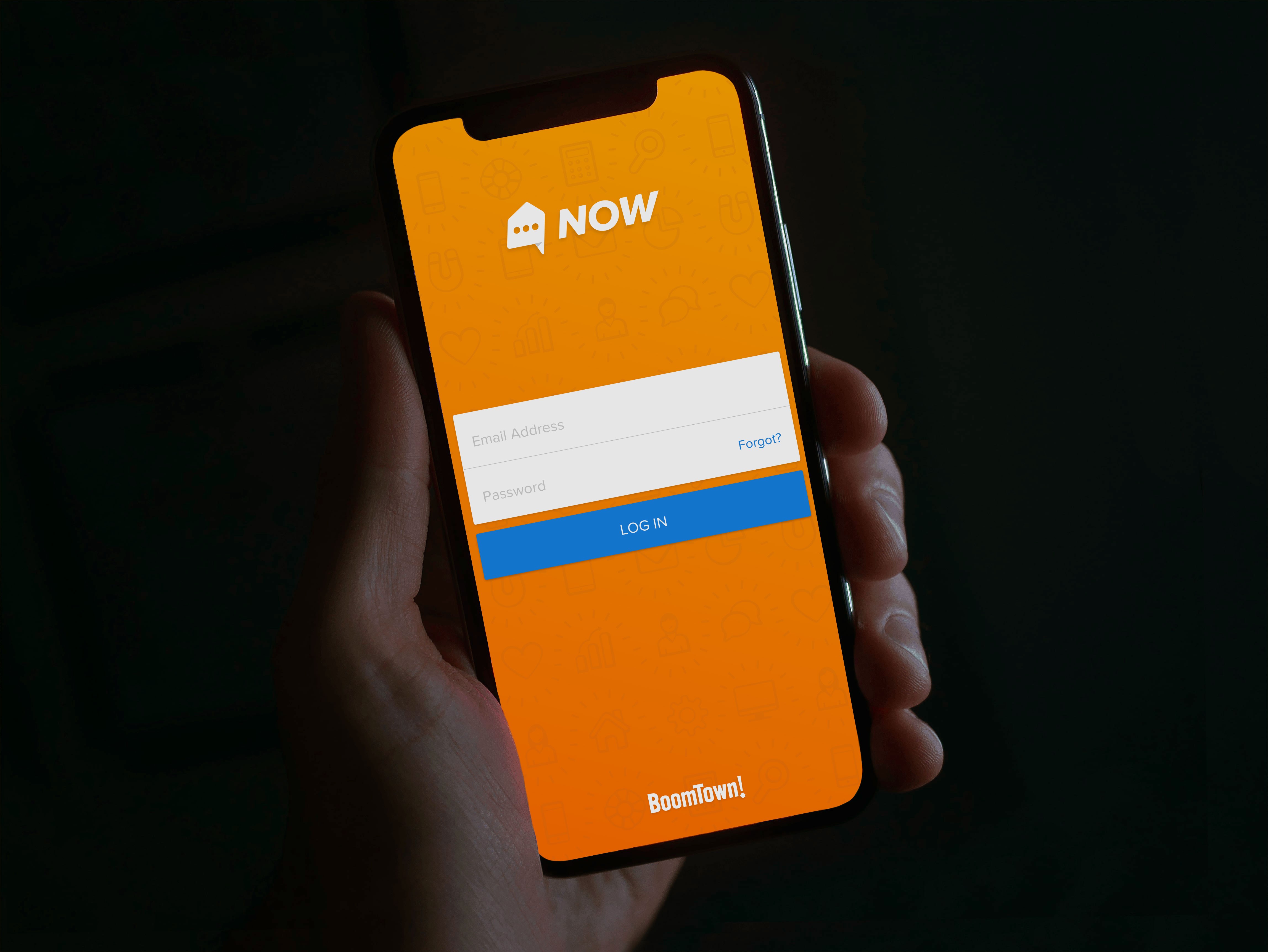 The NOW app login screen on a phone held in one hand, the orange brand screen with the NOW wordmark and BoomTown logo.